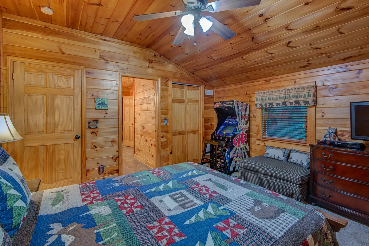 This inviting bedroom features a colorful quilt on the bed, warm wooden paneling, a classic arcade game machine, and a TV on the dresser. The setting balances cozy relaxation with playful entertainment.