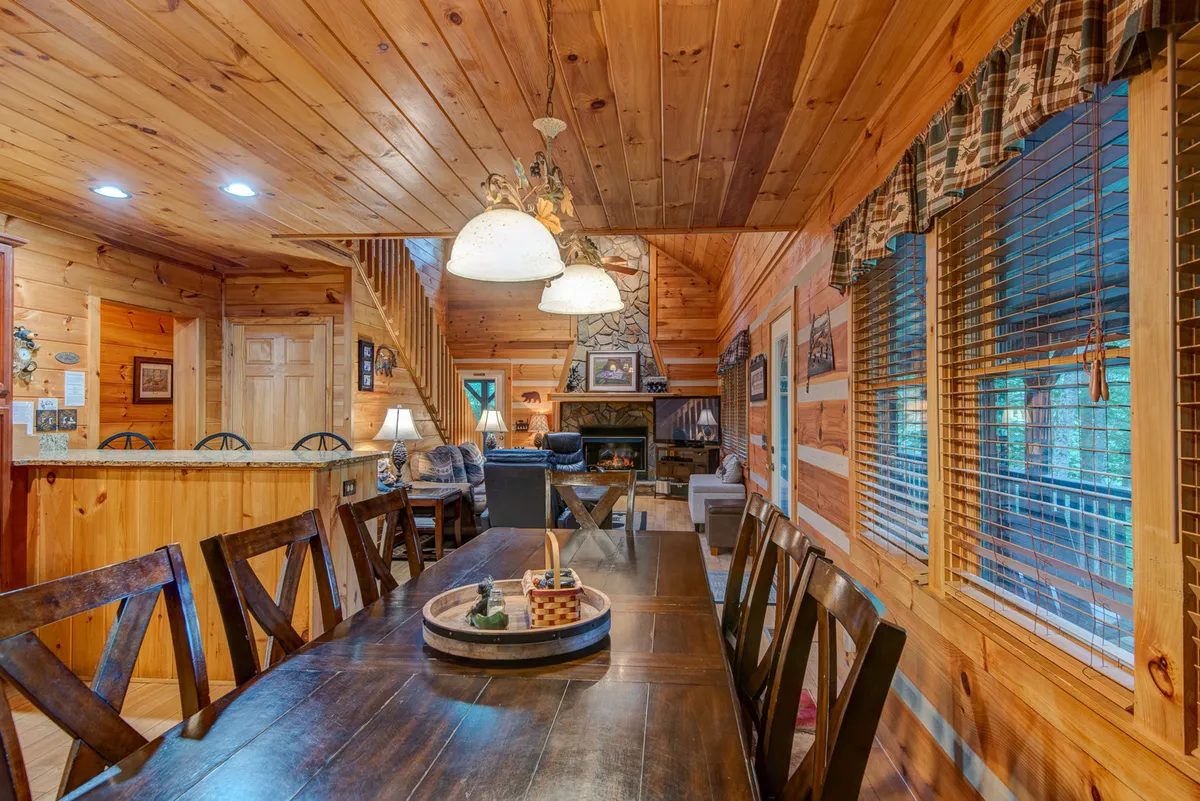 The image showcases a long wooden dining table under a chandelier, with a fireplace and staircase in view. A comfortable seating area is adjacent, highlighting a versatile, communal indoor space perfect for family dinners, celebrations, or game nights.