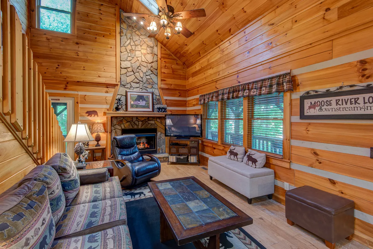 A warmly lit den featuring a grand stone fireplace, wooden beams, comfortable seating, and expansive windows that bathe the room in natural light. The arrangement of sofas and chairs encourages group gatherings and relaxation, while the stone hearth is the clear visual centerpiece.