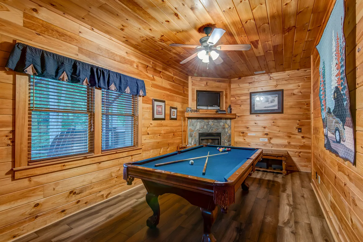 An inviting lower-level den with a pool table, wooden walls, a ceiling fan, and a fireplace. The space is designed for group games, friendly competition, and comfortable socializing.