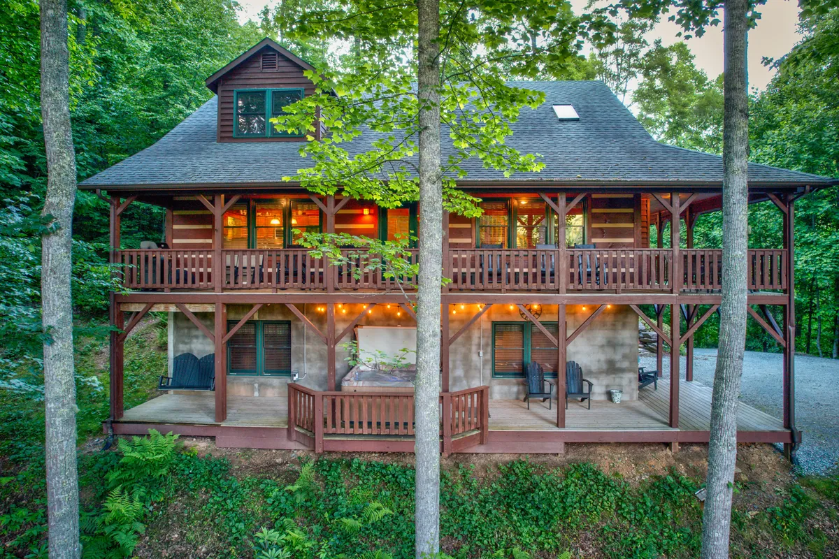 The image showcases a classic log cabin nestled among tall pines, featuring a spacious wraparound balcony with inviting outdoor seating, all bathed in the golden light of sunset. The tranquil setting hints at opportunities to relax with morning coffee while taking in mountain views, or gather with loved ones around a fire pit beneath the stars.