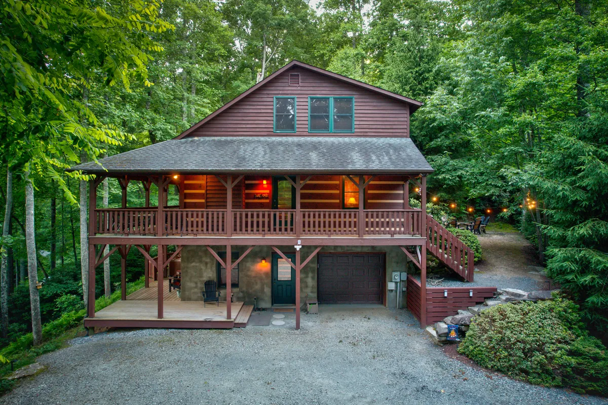 This image showcases the log cabin exterior surrounded by lush greenery, with a spacious porch area and visible driveway. The inviting porch offers outdoor seating, perfect for gathering before setting off to explore nearby attractions, while the natural setting hints at the region's adventure opportunities.