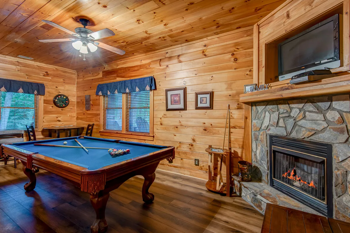 A lower-level recreation room with a pool table positioned beside a stone fireplace. Wooden walls, windows with blue curtains, and a relaxed seating area provide a welcoming setting for both lively games and quiet conversation.