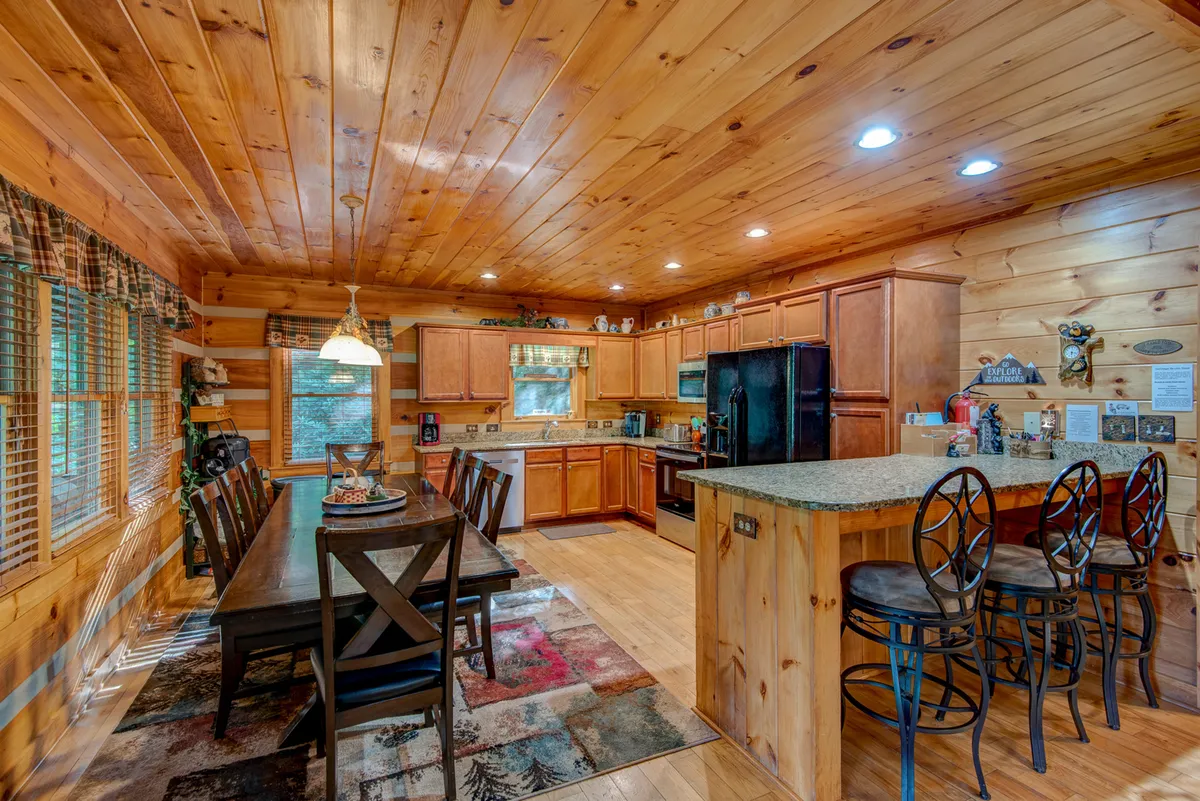 This image presents a spacious, wooden dining table paired with a bar area and stools, set amid kitchen cabinets and large windows. The setup is ideal for large group meals, casual gatherings, or festive celebrations, and the abundant natural light creates a welcoming, communal mood.