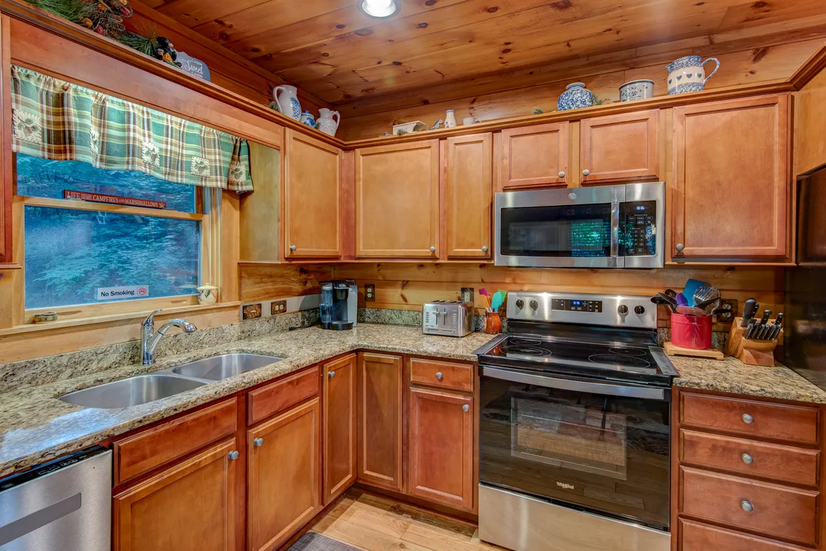 A close-up of the kitchen area featuring granite countertops, wood cabinetry, stainless steel appliances, and a window with a checkered curtain. The workspace is well-organized and inviting, suggesting ease of meal preparation for groups.