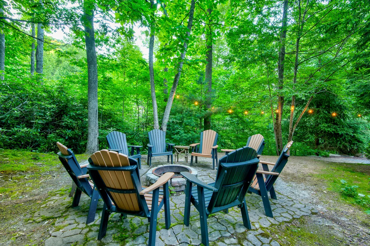 A cozy fire pit area encircled by Adirondack chairs, set among leafy trees. The setup encourages gathering for stories, laughter, and s’mores as dusk falls, with a strong sense of warmth and togetherness.