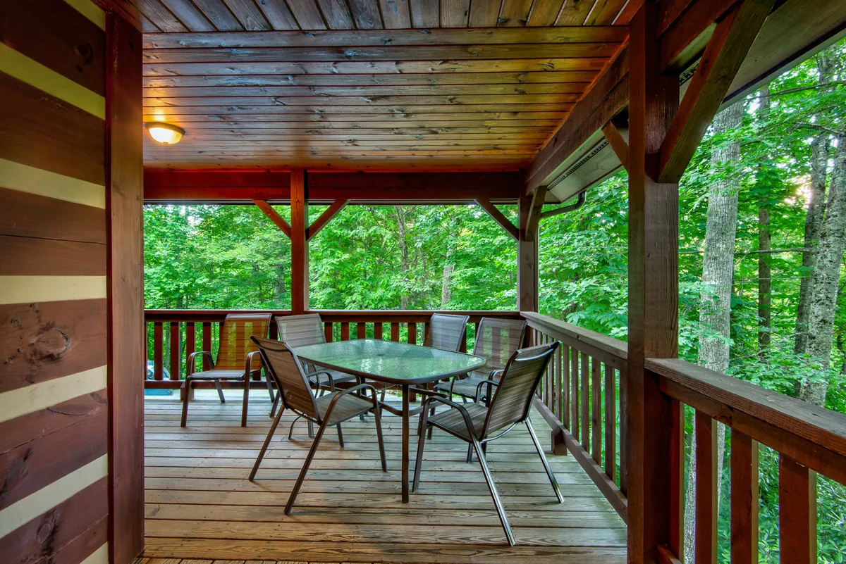 An exterior scene of a porch area with a glass table and metal chairs surrounded by lush greenery and a wooden railing. The setting is ideal for al fresco dining or morning coffee, with a view that connects the indoor dining experience to nature.