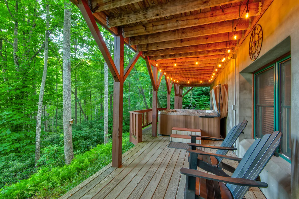 This vibrant exterior shot captures a spacious wooden deck with a hot tub, hanging string lights, and multiple rocking chairs. The deck is surrounded by lush trees, creating a private haven for relaxation, alfresco dining, or stargazing. The overall ambiance is welcoming and serene, with ample room for families and pets to gather.