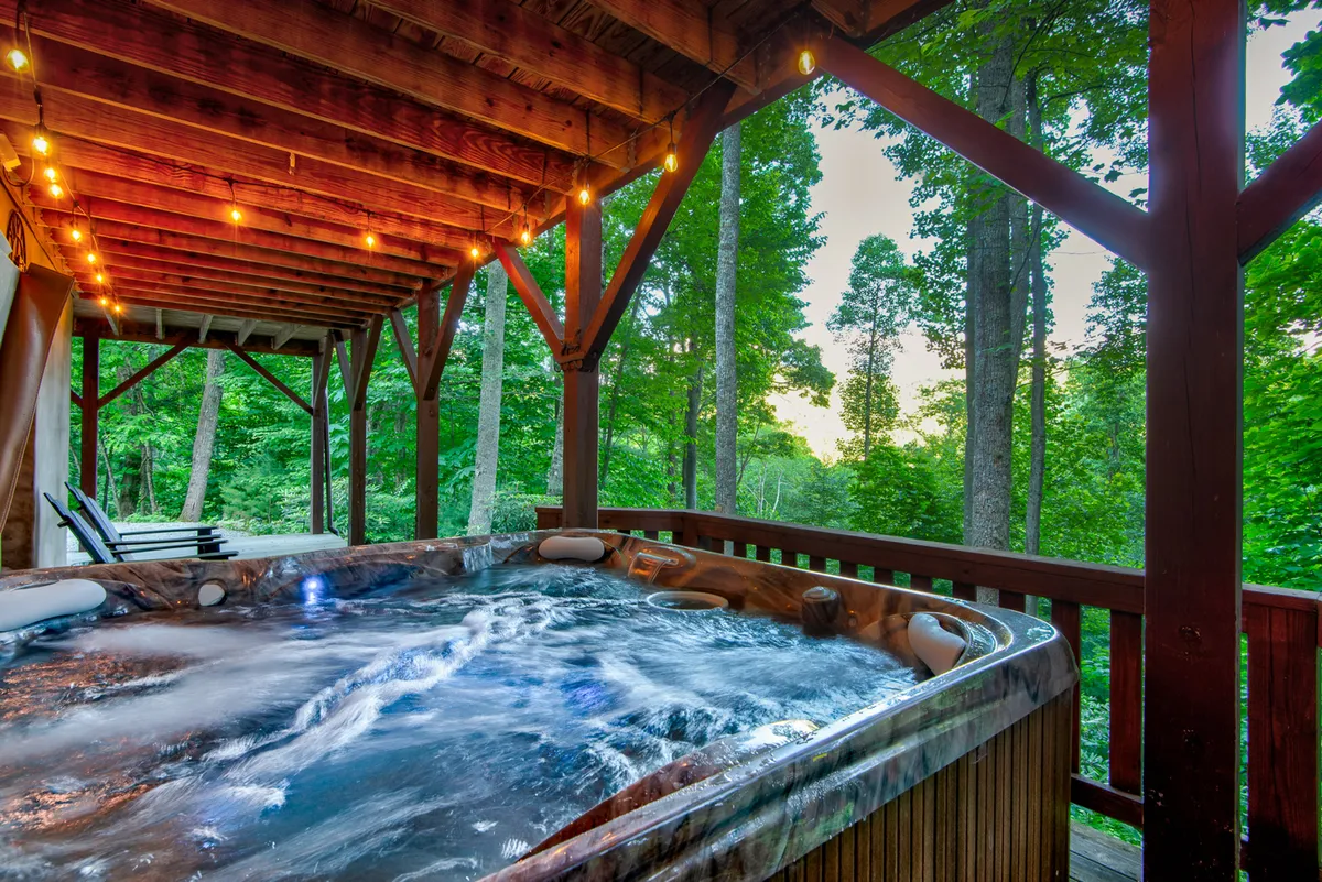 A tranquil wooden deck surrounded by trees, featuring a bubbling hot tub under string lights. The scene suggests relaxation and gathering, with natural beauty and privacy enhanced by the forest backdrop.